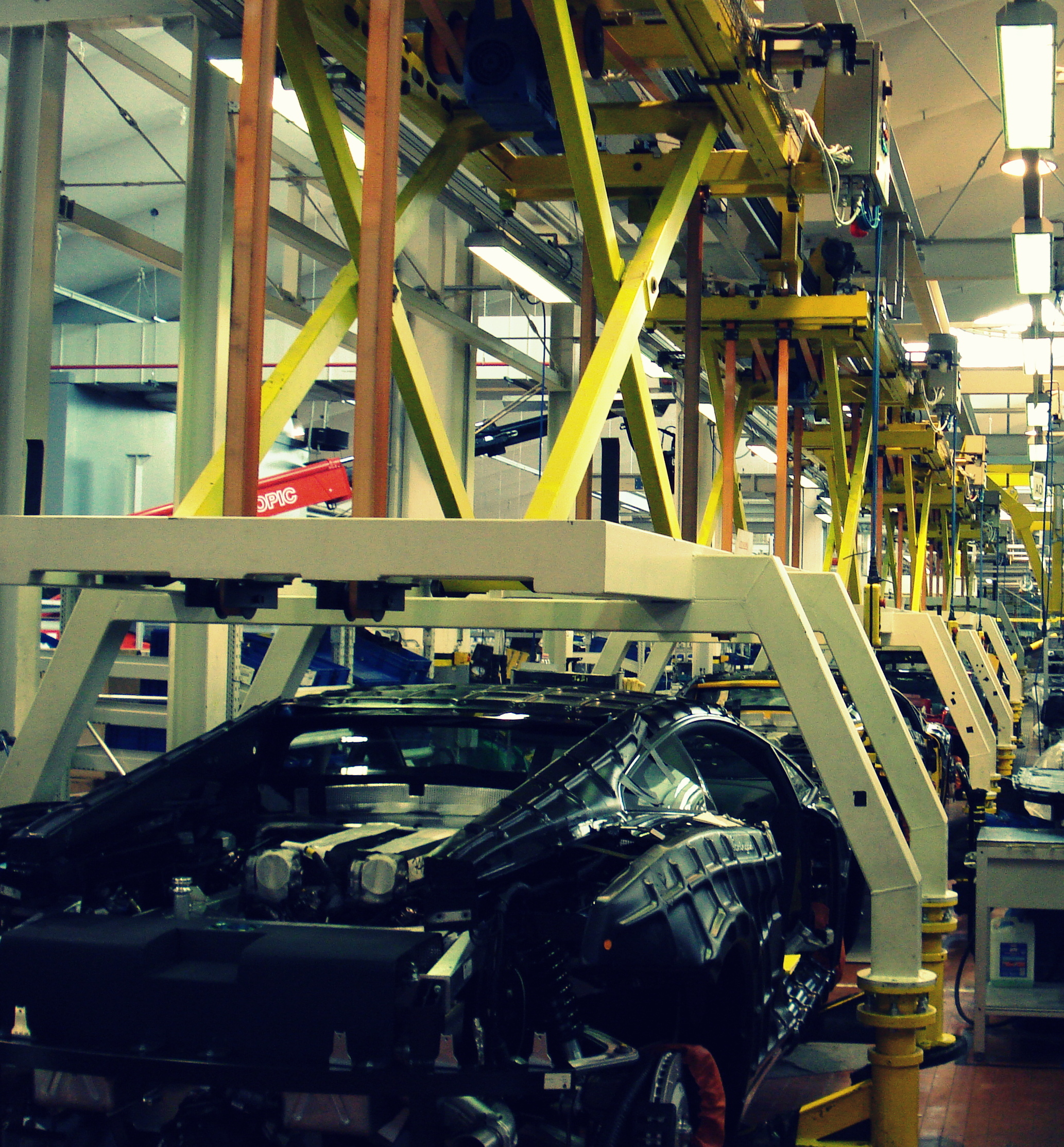 Linea di assemblaggio con più carrozzerie Lamborghini in avanzamento, trasportate da un sistema aereo giallo con strutture di supporto bianche.