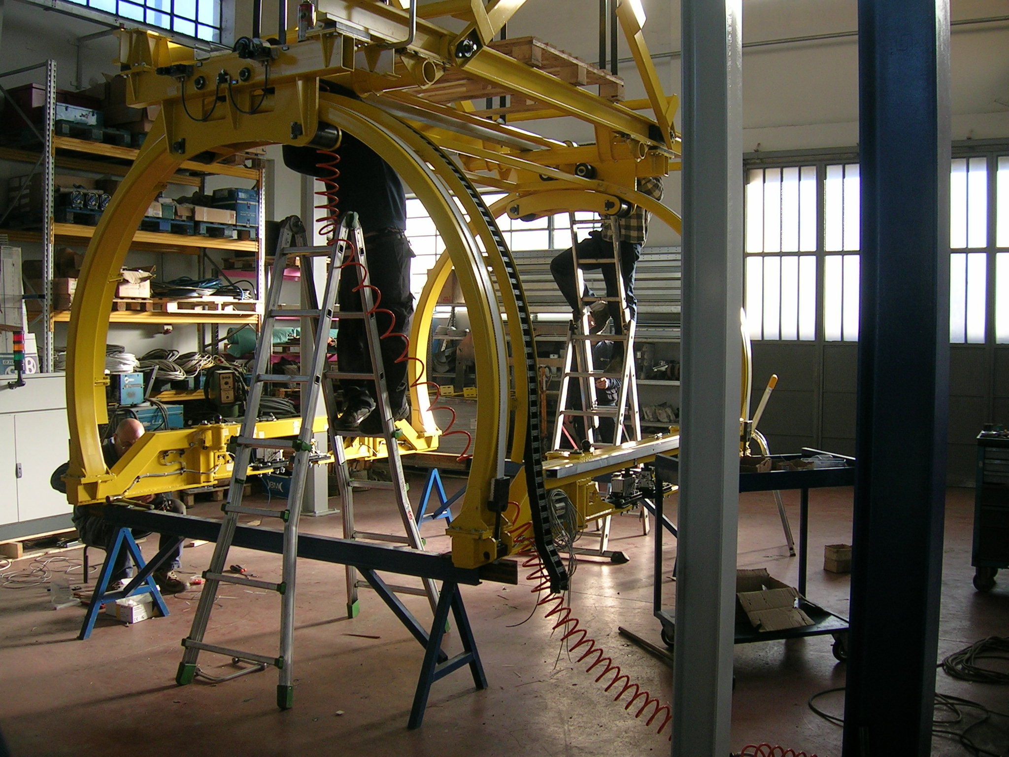 Tecnici al lavoro su una struttura gialla ad arco con scale e attrezzature per il montaggio di sistemi di sollevamento industriale.