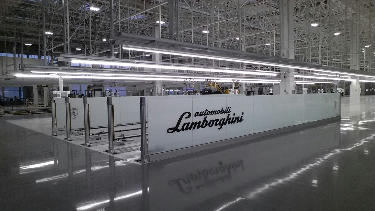 Impianto di trasporto Lamborghini con sistema su rotaie multiple, area delimitata da pannelli protettivi in vetro e design ottimizzato per produzioni di lusso.